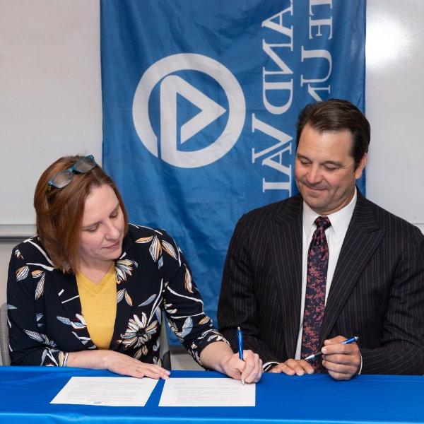 Two people are seated at a table with a blue table cloth. One person signs paper while another looks on. A Grand Valley State University flag hangs behind them.