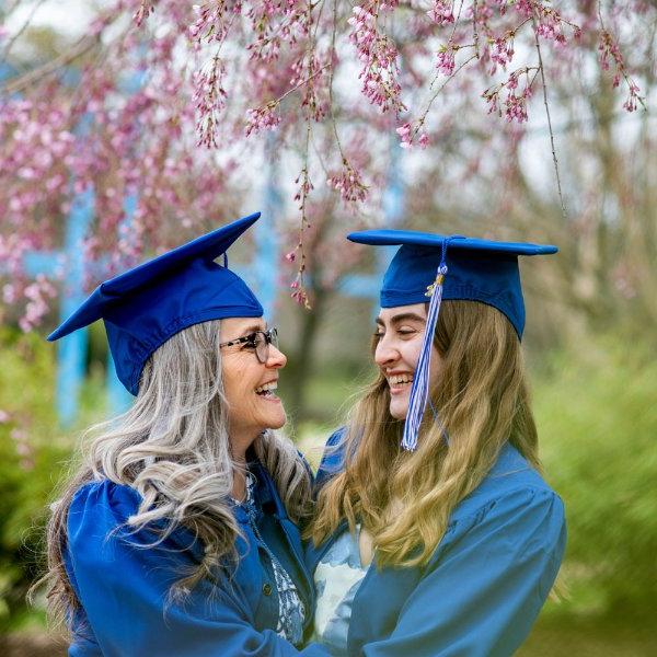 Two people wearing caps and gowns look at each other and are smiling. The blossoms from a tree hang overhead.